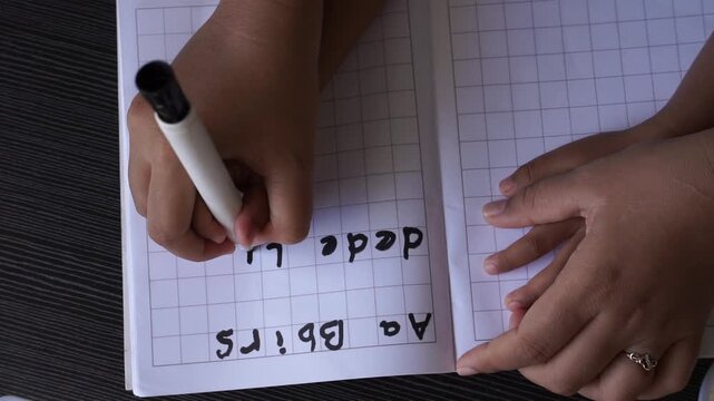 Overhead view of a mother guiding a child&rsquo;s hand using hand-over-hand technique while writing alphabet with a marker on a notebook, early learning and fine motor skill development