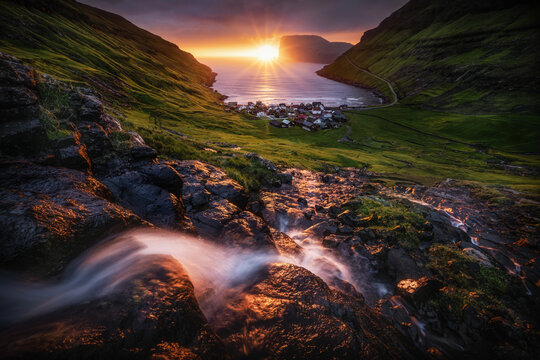 View of the village of Tjornuvik and a cascading waterfall on a rocky slope during a vibrant sunset over the Atlantic Ocean Tjornuvik, Streymoy, Faroe Islands.