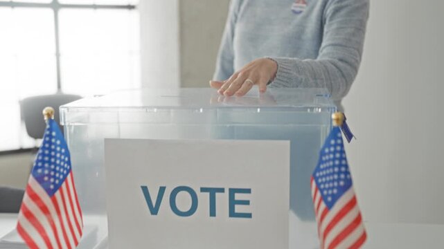 Woman inserting ballot into transparent ballot box on a table in a building polling station, flanked by small american flags; civic duty hopeful.