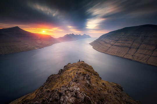 View of a hiker standing on the rocky summit of Klakkur overlooking the dramatic fjords and mountains during a vibrant sunset Klaksvik, Northern Isles, Faroe Islands.