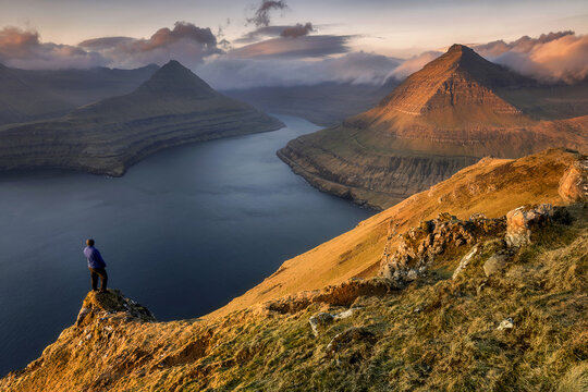 View of a person standing on a rocky cliff overlooking the deep blue fjord and pyramid-shaped mountains during golden hour in Funningur, Eysturoy, Faroe Islands.