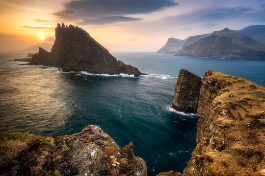 View of Drangarnir sea stacks featuring jagged rock formations and crashing waves under a golden sunset over the North Atlantic Ocean Faroe Islands.