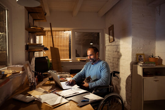 Man in wheelchair working from home office with cat at night