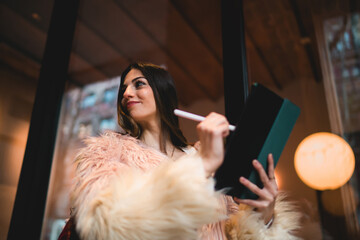 Happy young woman looking at camera holding stylus and tablet, successful female creator in faux fur coat enjoying creative process outdoors, authentic youth and digital lifestyle concept. © BullRun