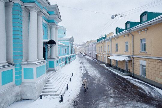 View of a person walking along a snowy street flanked by a bright blue neoclassical building with white columns and a yellow historic house in Moscow, Moscow, Russia.