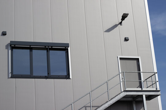 Modern industrial building exterior featuring a gray metal facade, large window, door, and metal staircase against a clear blue sky