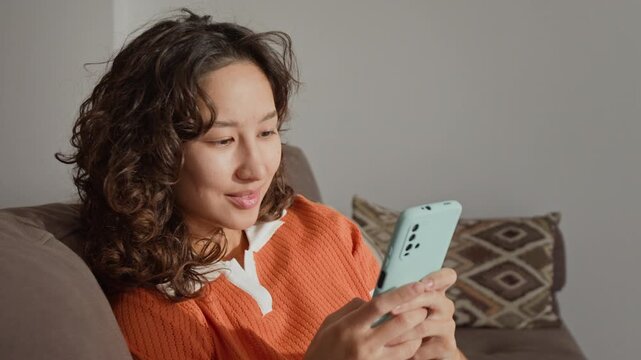 Asian young adult influencer sitting on sofa, using smartphone, scrolling social media feed and smiling. Footage suitable for mobile application advertising and social media marketing campaigns