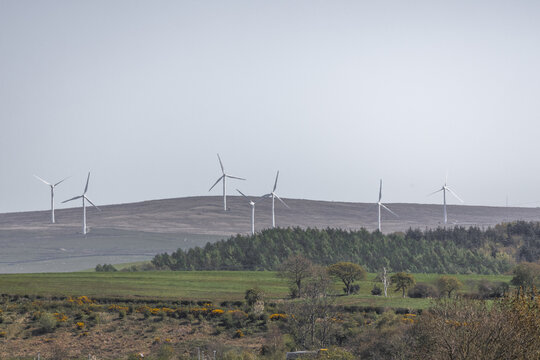 View of several wind turbines standing on a rolling hill above green fields and dense woodland under a clear sky Lancaster, England, United Kingdom.