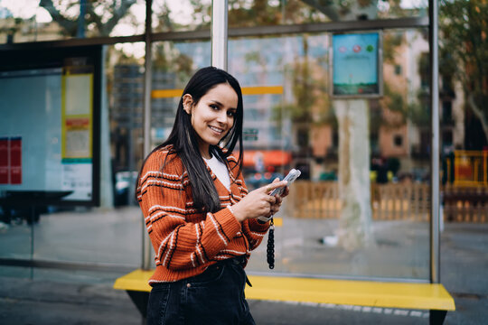 Latin woman standing at public transport stop checking phone, authentic representation inclusion equity real people, low angle shot urban environment, 5G internet connection.