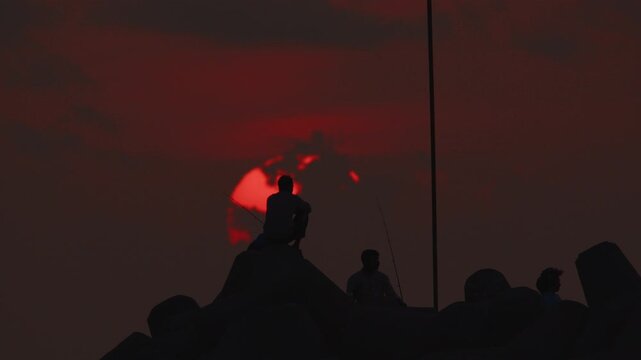 Telephoto close-up silhouette of a fisherman seated on a breakwater with the setting sun in the background.