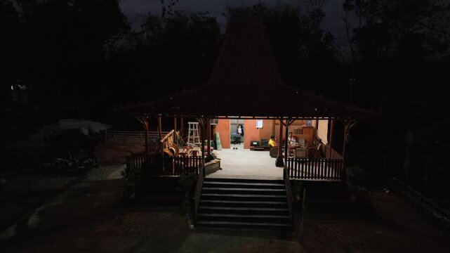 Nighttime rustic wooden cabin glows warmly. Cozy interior invites quiet reflection or rest. Outdoor seating faces a small, intimate porch space. Staircase leads to a softly lit entrance
