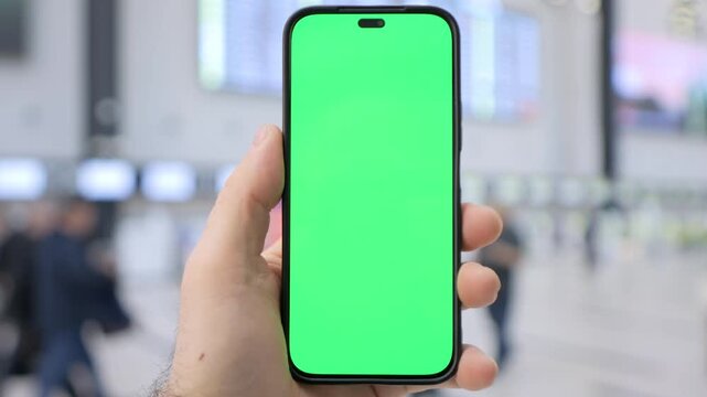 Hand holds phone with green screen in busy indoor space. Background shows blurred travelers moving past. Focus stays sharp on phone, emphasizing its blank screen. Ideal for mockups, app demos