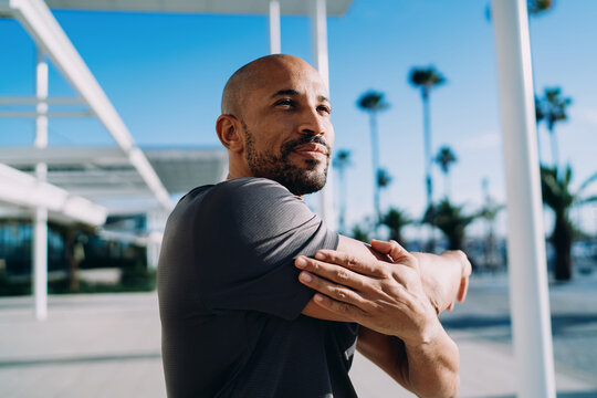 Low angle portrait of Black man stretching shoulder against blue sky and palm trees, focus on fitness and wellness, authentic representation, mindfulness, serenity, tropical city vibes.