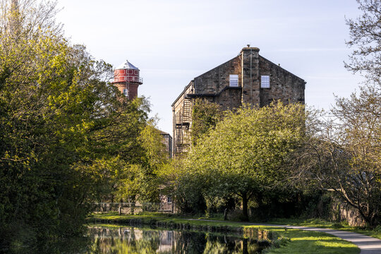 View of a historic stone building and a red water tower reflected in the canal surrounded by lush green trees in Lancaster, England, United Kingdom.