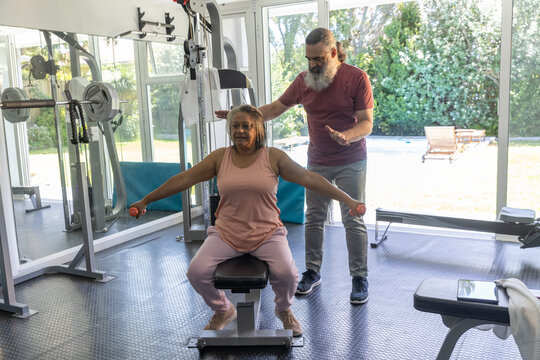 Senior African American woman in pink doing lateral raises on bench with weights, trainer guiding