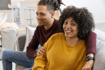 Fototapeta premium African American couple sitting on light sofa in bright living room wearing sweaters and smartwatch