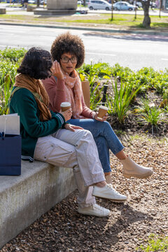 Two female friends sitting on low bench at streetside holding takeaway coffee cups with navy bag