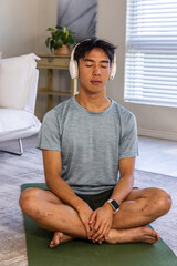 Obraz premium Asian man meditating on green yoga mat in living room, wearing tee, shorts, headphones, smartwatch