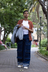 African woman walking on paved park path holding gray sweatshirt, wearing glasses and sneakers