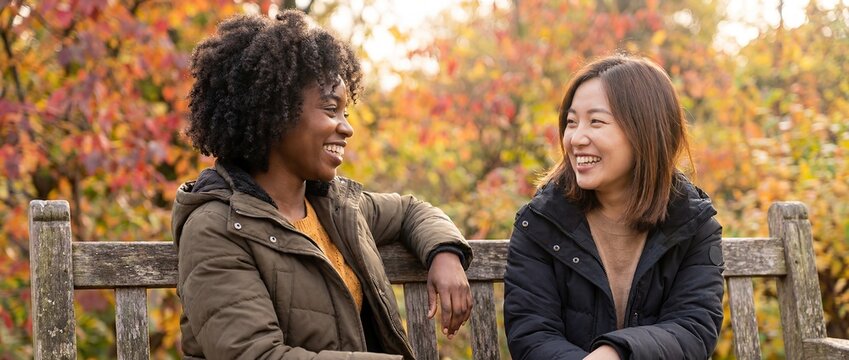 interracial female friendship Black East Asian friends talking autumn outdoors women face each other during relaxed conversation on park bench showing attentive listening mutual respect and cultural