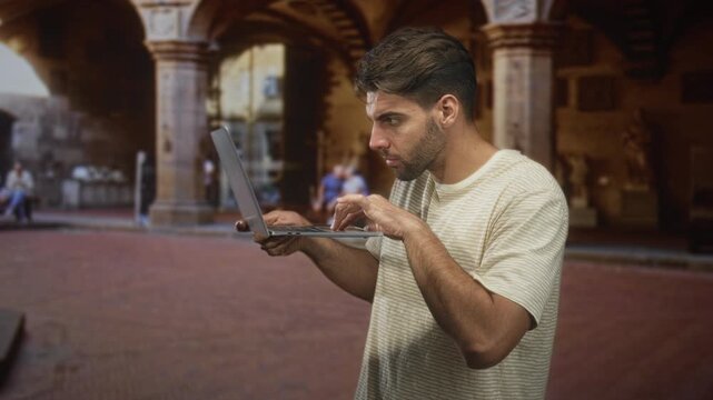 Man typing on laptop while holding it at chest level in a historic building arcade plaza with columns and arches, intent expression and upright posture; concentration productivity.