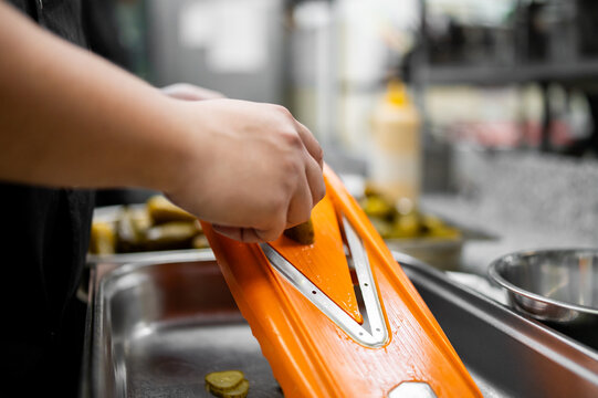A chef using an orange mandoline slicer to slice pickles into a metal tray in a commercial kitchen.