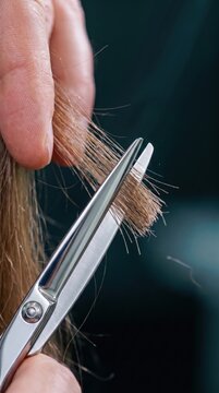 Macro view of fingers holding blonde hair ends while sharp metal scissors trim the strands against a dark blurred background.
