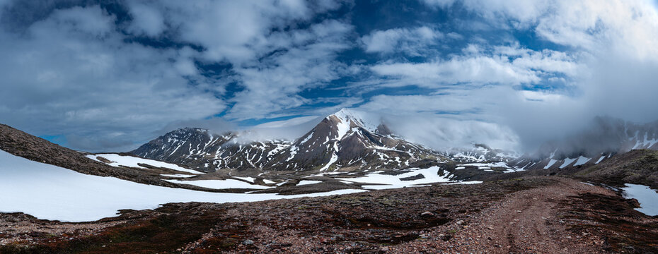 View of Brunavik mountains with snow-covered peaks, rugged rocky terrain, and a winding dirt trail under a cloudy blue sky Bakkagerdi, Mulathing, Iceland.