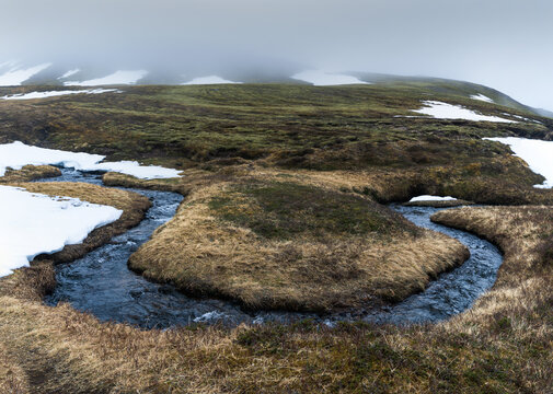 View of the winding stream in Brunavik with patches of melting snow and mossy tundra under a foggy sky in Bakkagerdi, Mulathing, Iceland.