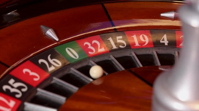A roulette wheel rotates around its central spindle while a white ball rests in pocket 0. The numbered pockets form a continuous ring divided by metal separators on a polished wooden track. 