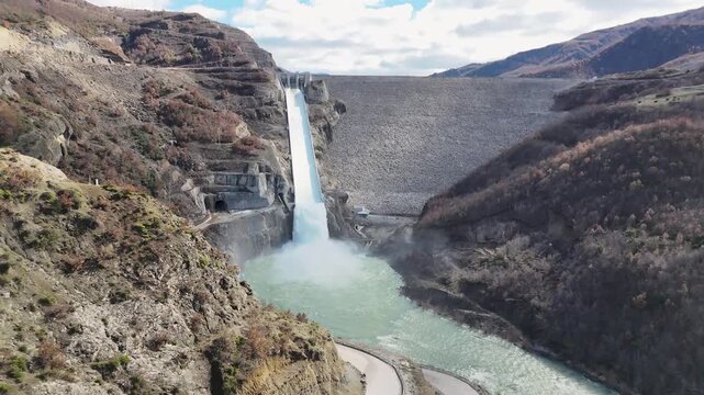 Aerial drone camera moving forward and tilting down over the massive Moglic&euml; Dam spillway in Albania, capturing rushing white water plunging into the mountain river canyon