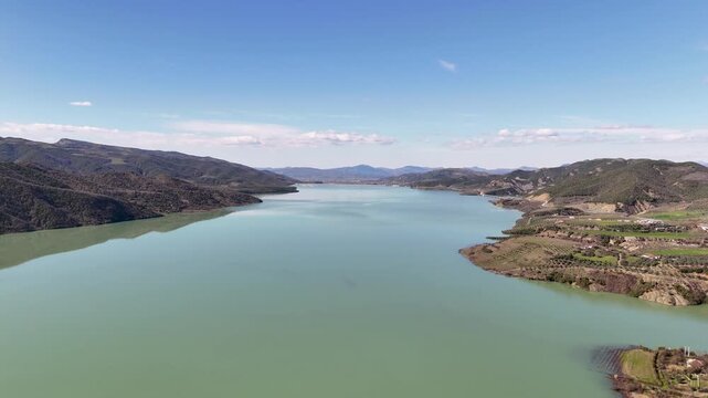 Aerial drone camera moving forward high over the sprawling Banja Reservoir in Albania, capturing the vast pale green water, rugged shorelines, and distant rolling hills