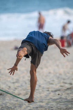 View of a man balancing on a slackline on a sandy beach with ocean waves and blurred figures in the background Rio de Janeiro, State of Rio de Janeiro, Brazil.