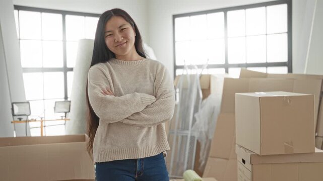 Woman with crossed arms beside stacked cardboard boxes and packing materials in a sunlit building; moving day contentment.
