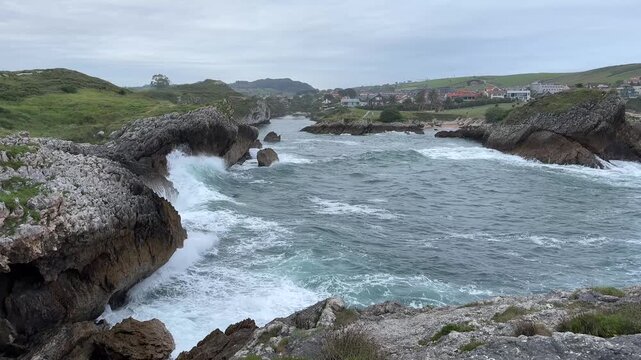 Natural Atlantic water movement within one of the rocky bays of Costa Quebrada, with weathered cliffs, coastal village and cold gray ocean in northern Spain.
