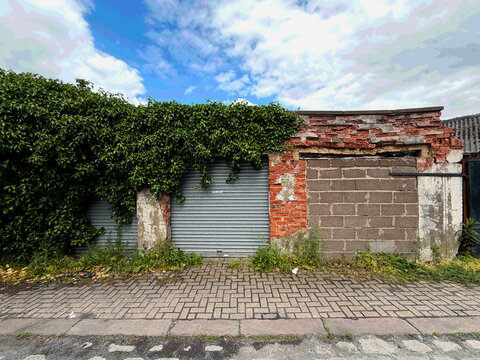 A weathered metal doored garage, its red brickwork crumbling beneath a veil of lush green ivy, sits under a bright, cloud‑washed sky on Paternoster Lane, Great Horton, Bradford, UK.