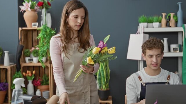 Woman arranging tulip bouquet with hands while man uses laptop in a flower shop building; collaboration teamwork.