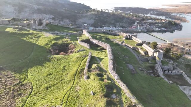 Aerial camera circles counterclockwise over the ancient stone walls and green grassy courtyards of Rozafa Castle in Shkod&euml;r, Albania, during a bright morning sunrise