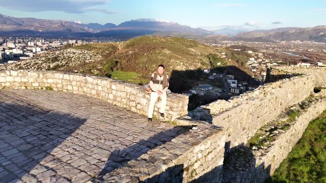 Aerial drone camera pulling back and rising from host Charles Huang sitting on a stone wall at Rozafa Castle in Shkod&euml;r, Albania, revealing the historic fortress and sunlit valley