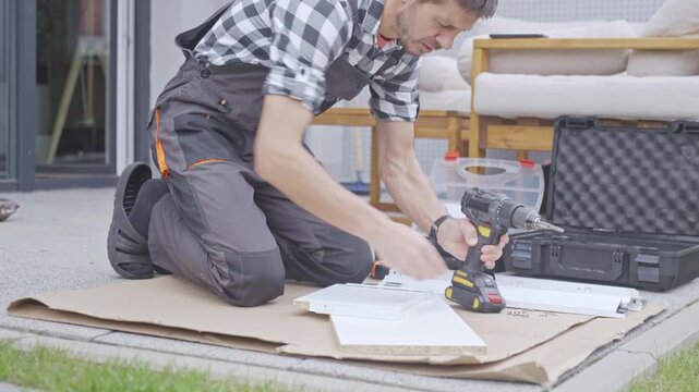 Man in work overalls takes screw and starts fastening furniture panel on cardboard outside