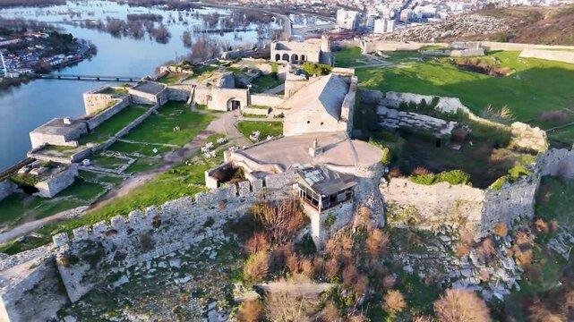 Aerial circles the historic stone walls, ruins, and courtyard buildings of Rozafa Castle in Shkod&euml;r, Albania, illuminated by warm sunset light with the Buna River in the background