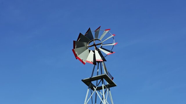 A windmill stands tall in a rural area, turning gently in the wind against a clear blue sky. The structure is designed to capture wind energy for practical applications.