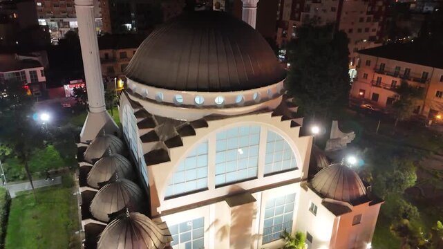 Night drone camera rising vertically while panning down over the Ebu Bekr Mosque in Shkod&euml;r, Albania, framing its large illuminated dome, stepped facade, and minaret