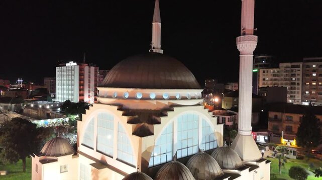 Aerial descends vertically behind the illuminated main dome of Ebu Beker Mosque, framing its twin white minarets and the glowing night cityscape of Shkod&euml;r, Albania