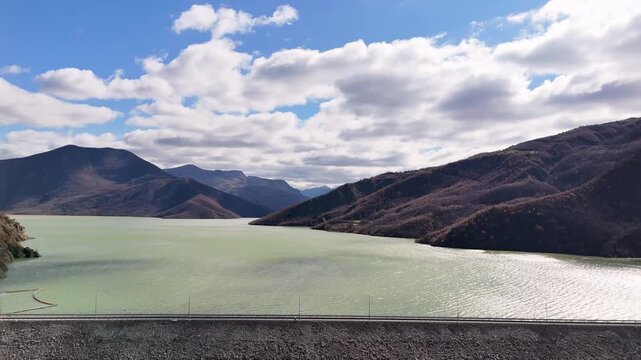 Aerial drone camera pulling back over the massive rockfill Moglic&euml; Dam wall in Albania, revealing the vast green reservoir lake nestled within sunlit mountain ridges under a cloudy blue sky
