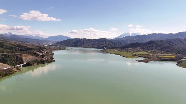 Aerial drone camera moving forward high over Lake Banja reservoir in Albania, revealing winding valley roads, green agricultural plots, and distant snow-capped peaks