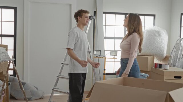 Woman hugging man amid unpacked cardboard boxes, bubble wrap, rolled rug and ladder in a sunlit living room; homecoming happiness togetherness.