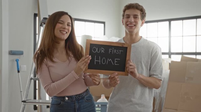 Woman and man holding a chalkboard that reads our first home in a building, smiling at camera; new home joy.