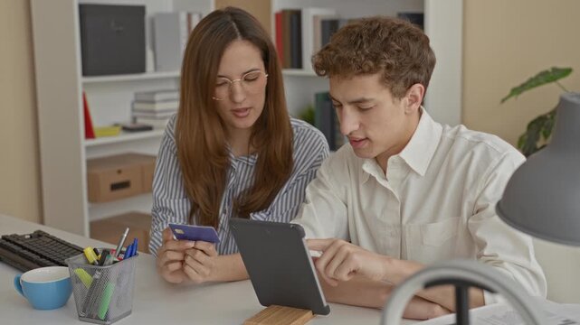 Man and woman sit at desk hold creditcard and man points to tablet on desk inside office building; financial planning trust.