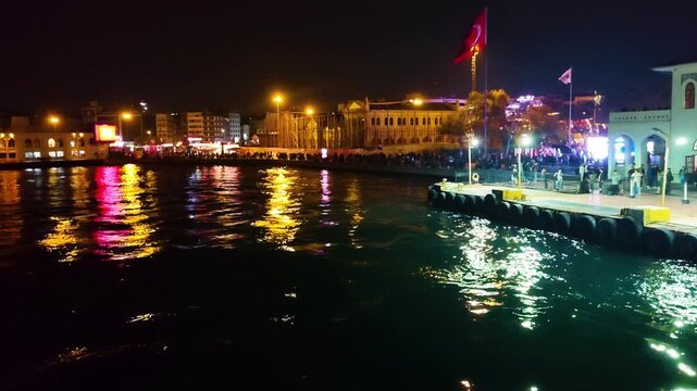 View from an approaching passenger boat to the historic Kadikoy ferry pier at night with people waiting, city lights reflecting on sea water and a large waving Turkish flag in Istanbul, Turkey